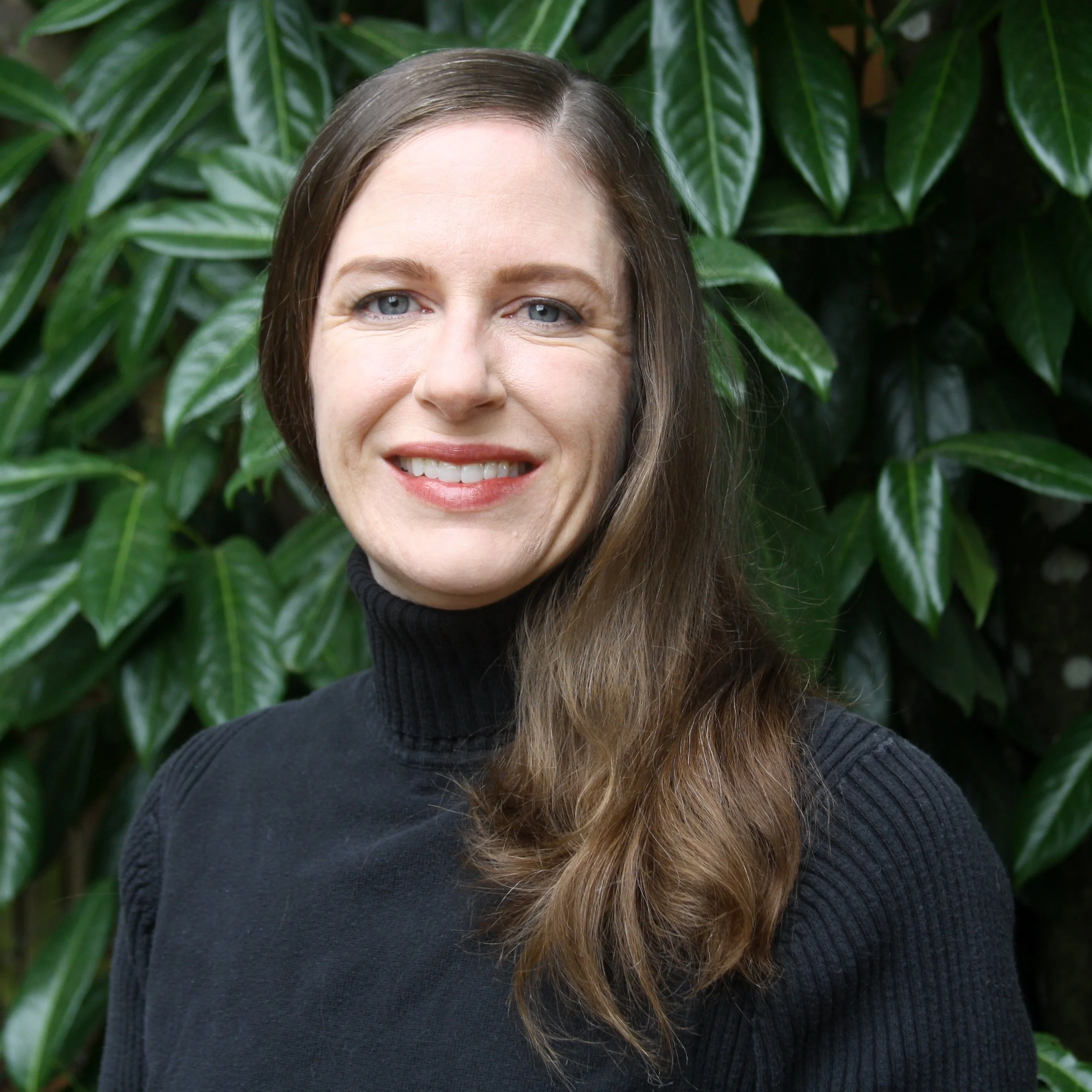 A smiling white woman with shoulder-length wavy brown hair smiles at the camera against a backdrop of dark green leaves; she is wearing a black turtleneck sweater.