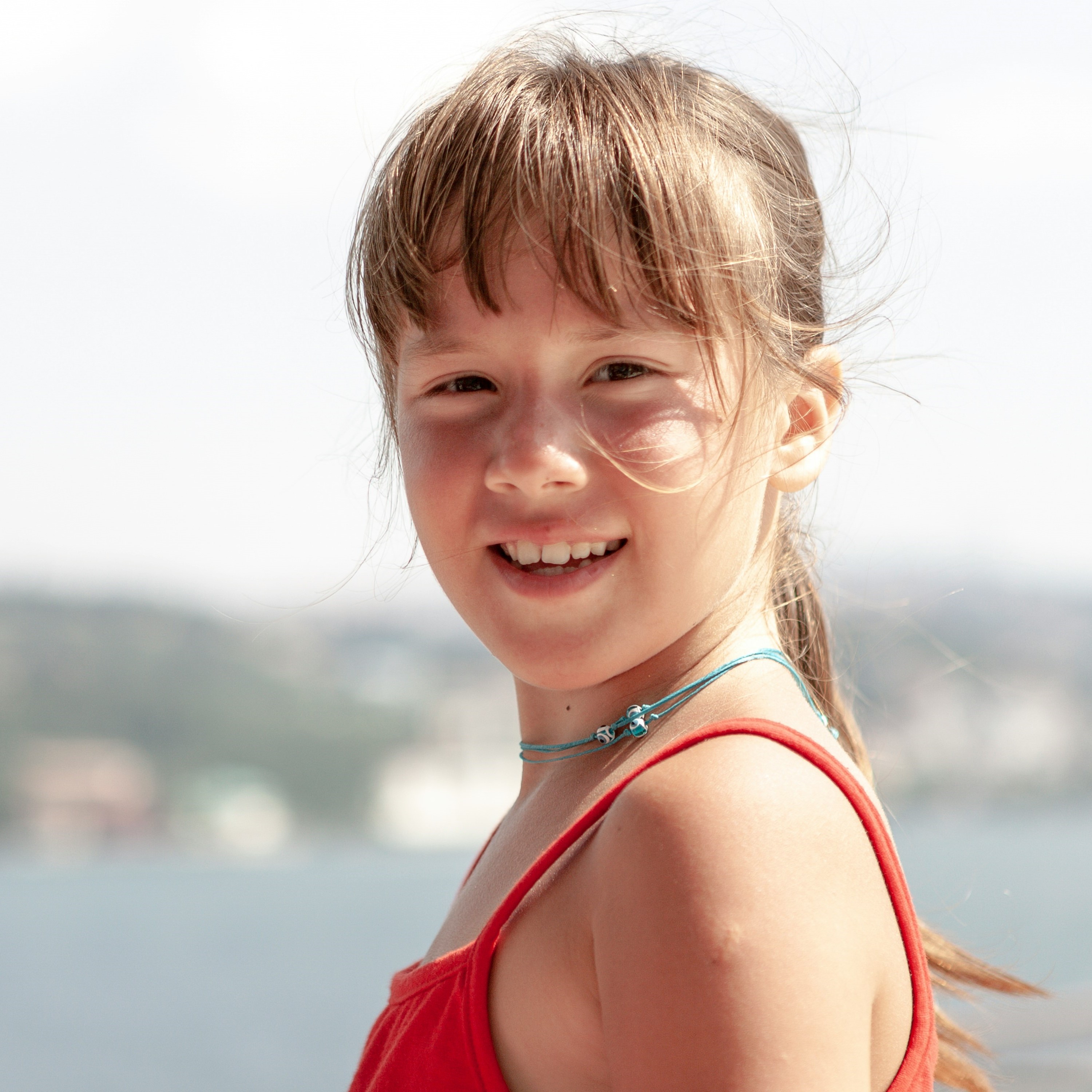 A head and shoulders image of a smiling girl with tanned skin and damp, windblown hair; the sun is shining on her and she is wearing a necklace made of blue cord.