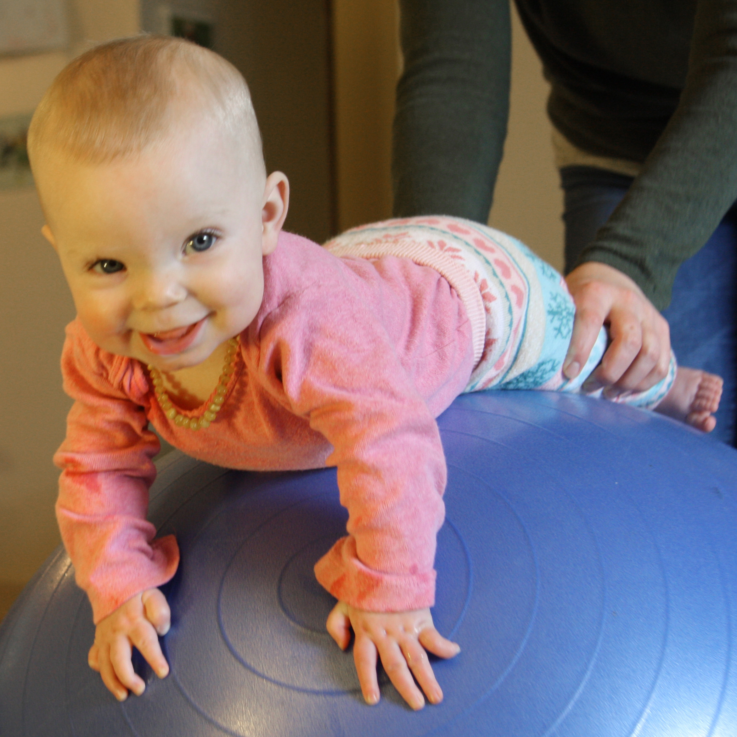 A smiling white baby in a pink shirt plays tummy-down on a yoga ball while having their legs supported by their parent in the background. Photo by Elizabeth Ryan